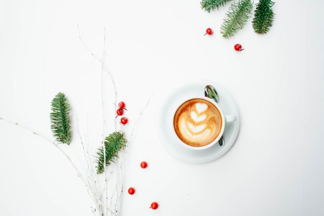 A festive holiday scene features a cup of coffee with latte art surrounded by evergreen branches and red berries on a white background. Perfect for promoting holiday-themed events, coffee shop menus, and seasonal advertisements. The image exudes a cozy winter feel and can be used in social media posts, holiday greeting cards, or cafe promotions.