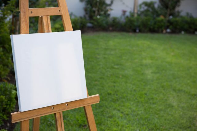 Blank canvas on an easel set in a lush garden with green grass. Perfect for themes related to art, creativity, and outdoor activities. Ideal for illustrating artistic inspiration, hobby pursuits, or promoting art supplies and outdoor painting workshops.