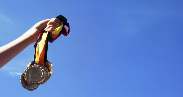 A hand is raised against a clear blue sky, holding a collection of medals, with copy space. It signifies achievement and success in competitive events, in sports or academic fields.