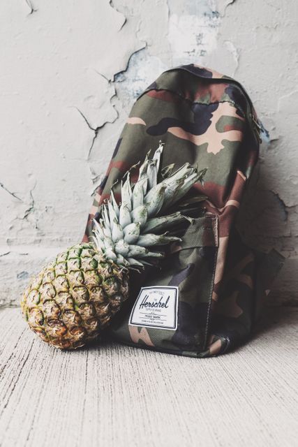 Camo backpack placed on ground against a weathered wall with a fresh pineapple beside it. The blend of camo pattern with natural pineapple creates a visually intriguing contrast of urban and natural elements. Perfect for use in fashion, lifestyle, and travel content emphasizing trendy urban styles and outdoor adventure.