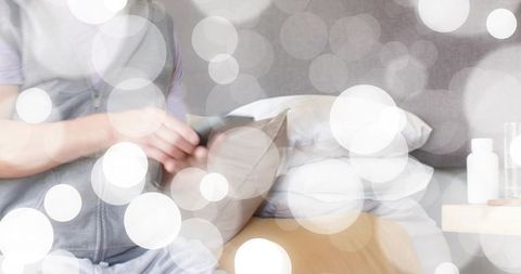 Asian man holding throw pillow sitting bedside in grey hoodie with cozy bokeh light