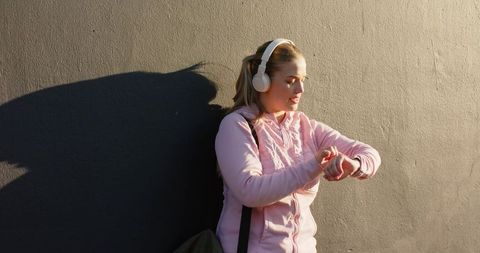Young woman leaning on sunlit wall checking smartwatch while listening to music outdoors