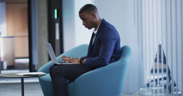 African American businessman sitting on modern chair in stylish office, working on laptop with intense focus. Great for articles, advertisements, or visuals highlighting professional life, remote work, business technology, and productivity. Ideal for corporate websites, business blogs, and workplace wellness content.