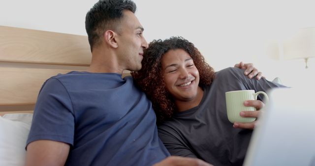 Happy diverse gay male couple on bed, having coffee and using laptop in the morning. Communication, free time, gay, relationship, togetherness, domestic life and lifestyle, unaltered.