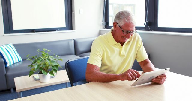 Elderly man with glasses using a tablet in a bright office space. Highlights senior engagement with technology. Useful for topics related to digital literacy, aging population, office design, and senior-friendly technology solutions.