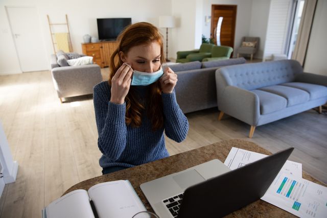 Woman wearing face mask working on laptop at home, emphasizing remote work and safety during the COVID-19 pandemic. Useful for illustrating concepts of social distancing, health precautions, and the shift to home office environments.