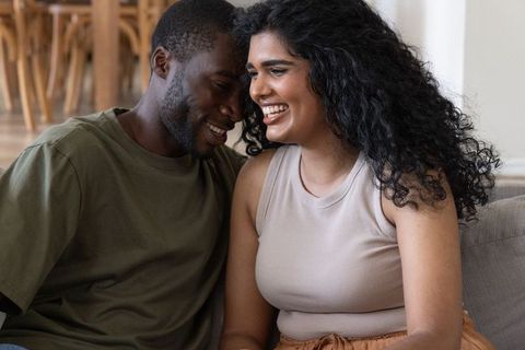Interracial couple laughing and leaning on sofa, sharing intimate cozy home moment