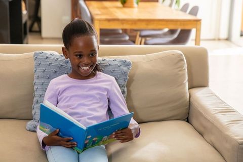 African american girl reading on sofa, smiling during cozy sunlit storytime at home