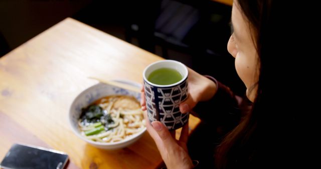 This image captures a woman enjoying a traditional Japanese meal of udon noodles accompanied by a cup of green tea in a casual restaurant. Suitable for use in articles about Japanese cuisine, dining experiences, food blogs, restaurant promotions, or cultural commentary.