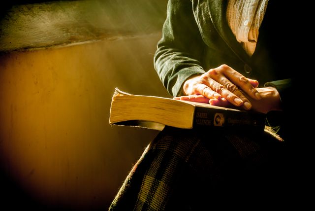 Senior person resting hands on an old book bathed in soft, golden light from nearby window. Showing a quiet, contemplative moment, highlighting importance of reading and wisdom. Perfect for themes related to aging, knowledge transfer, education, and peaceful living.