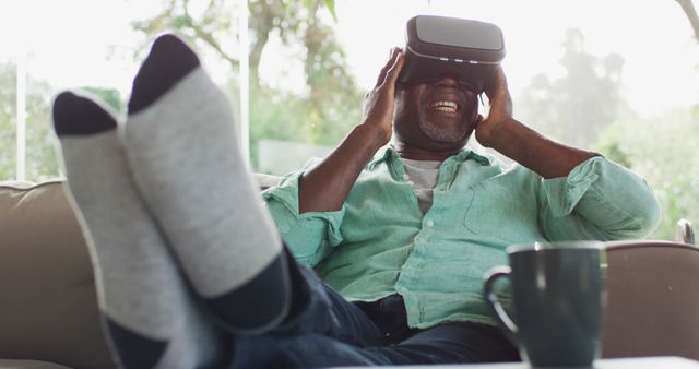 Happy african american senior man sitting with feet up using vr headset and laughing. retirement lifestyle, spending time alone at home.