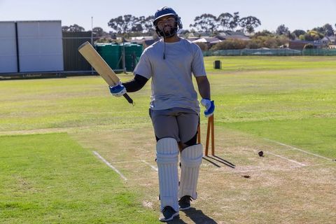 Cricketer walking off pitch raising bat after innings on sunny suburban ground