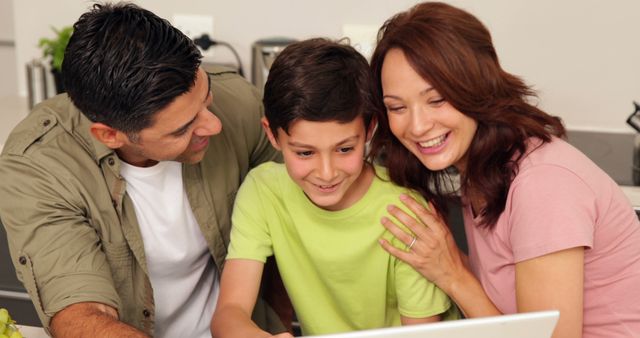 This image shows a cheerful family of three smiling and using a laptop together at the kitchen table. The parents and child are closely gathered, indicating a moment of bonding and connection. The setting conveys warmth, togetherness, and modern family life. This image is ideal for use in advertisements, articles, and promotional materials highlighting family-friendly technology, internet safety, digital learning, and family activities.