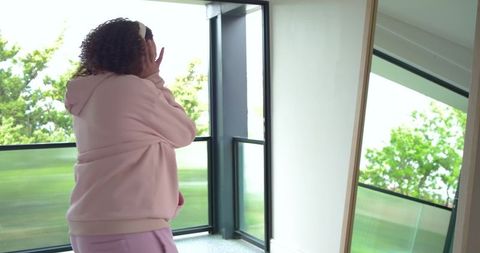 African american woman wearing pink hoodie looking out through glass balcony door
