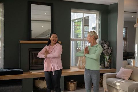 Mature women practicing namaste yoga together in cozy contemporary living room
