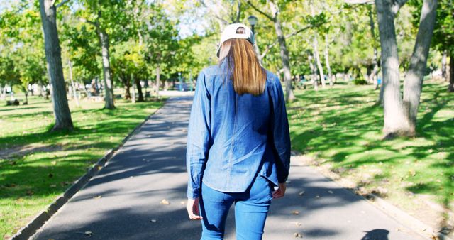 Back view of biracial woman with cap walking down alley in sunny park. Free time, lifestyle, relaxation and city break, unaltered.