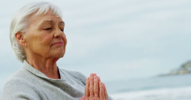 A senior Caucasian woman practices meditation or prayer by the beach, with copy space. Her serene expression suggests a moment of peace or reflection in a tranquil outdoor setting.