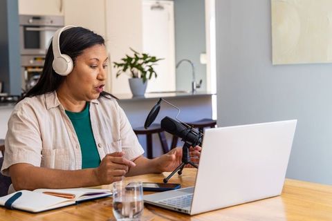 Mature woman recording podcast at home kitchen table with laptop and microphone