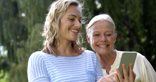 Two women are smiling and looking at a smartphone outdoors, enjoying time together. This image can illustrate concepts such as family bonding, intergenerational relationships, technology usage across different age groups, and sharing special moments. Suitable for articles, advertisements, or presentations on family dynamics, technology in daily life, or outdoor leisure activities.
