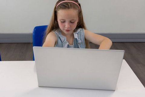 Young girl studying with laptop at desk in minimalist classroom, focused calm learning