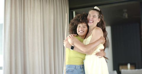 Two friends hugging and laughing in sunlit living room holding pool cue and smartwatch