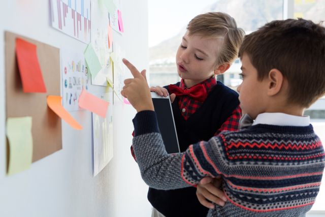Two children dressed as business executives are discussing ideas in an office setting, pointing at a bulletin board filled with notes and charts. This image can be used to represent concepts of early education, business training for kids, teamwork, and innovative learning environments.