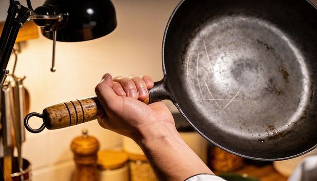 Hand gripping seasoned cast iron skillet with wooden handle, lamp and pepper mill