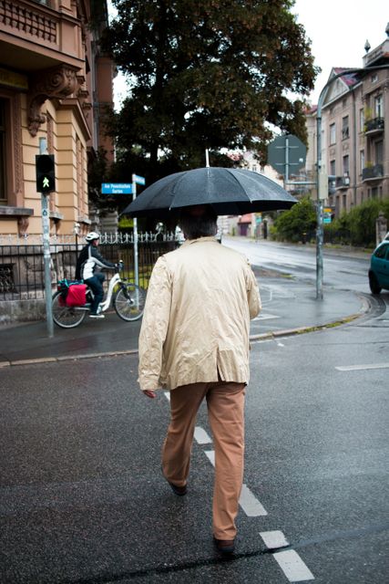 Person carrying a black umbrella walking on wet pavement during rain in urban setting. Suggested uses: transportation services, urban planning, weather-themed publications, rainwear fashion advertisements.