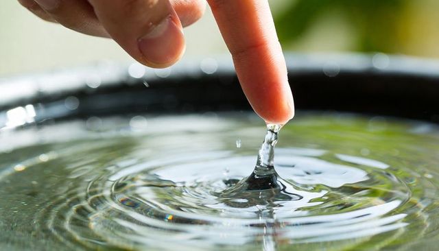 Finger touching water creating glassy droplet and concentric ripples in sunlit basin