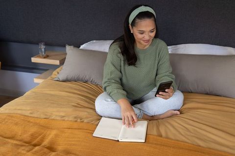 Mid-adult asian woman relaxing on bed browsing smartphone and journaling in cozy bedroom