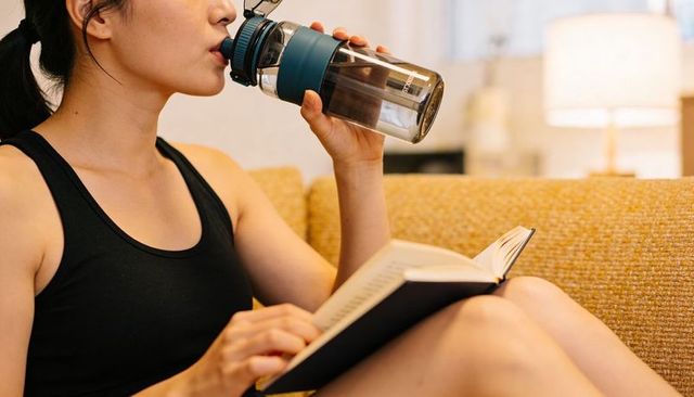 Asian woman reading and drinking from reusable bottle while relaxing on cozy sofa at home