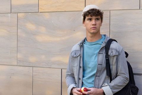 Male teen in beanie standing by sunlit urban stone wall holding bitten apple, casual