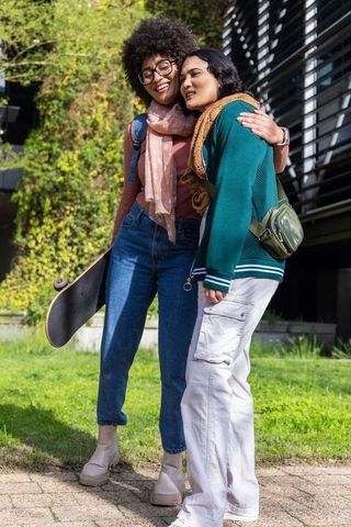 Diverse young women embracing on campus with skateboard and backpacks, casual friendship