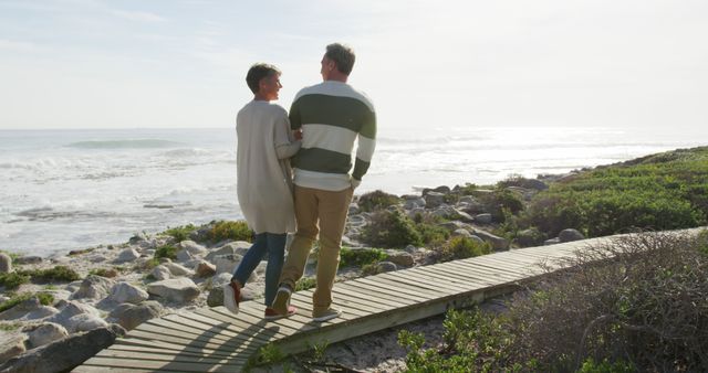 Middle-aged couple walking arm-in-arm along a scenic beach boardwalk next to the ocean. Perfect for advertising retirement, travel destinations, lifestyle blogs targeting older couples, or illustrating concepts of love, companionship, and relaxation in a natural environment.