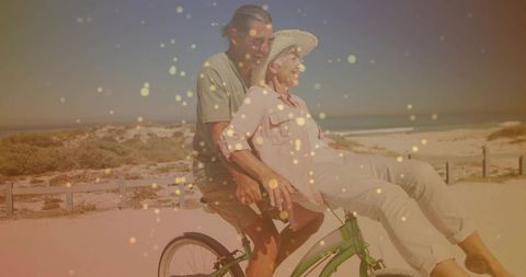 Senior couple riding green cruiser bicycle along sunlit sandy beach with golden bokeh