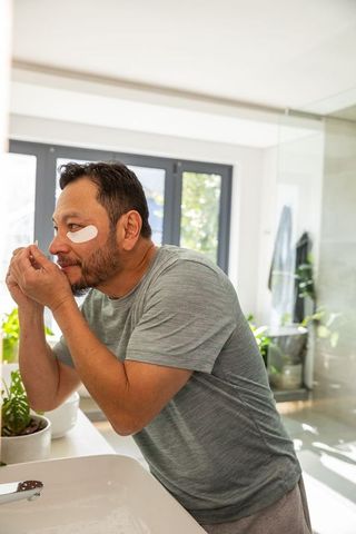 Mature asian man applying eye patches during morning skincare routine in bright bathroom