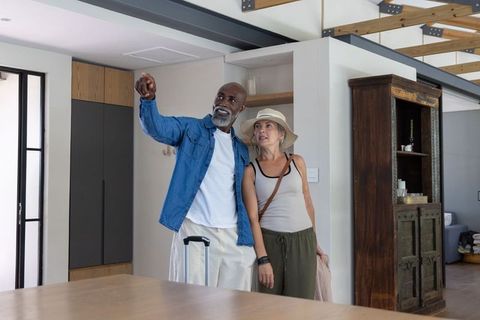 Mature couple touring modern rental home with luggage, pointing at interior features