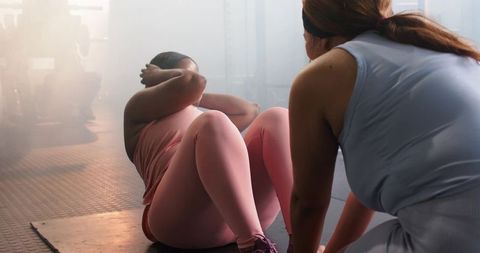 Diverse female trainer coaching client performing sit-ups on mat in backlit gym