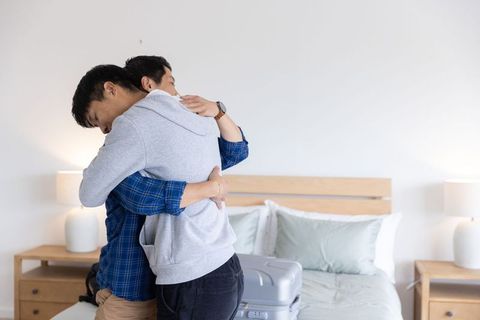Two men sharing warm goodbye hug in minimalist bedroom with silver suitcase on bed