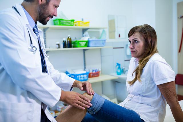 Male doctor examining female patient's knee in a hospital room. The doctor is focused on the patient's knee, likely assessing for injury or pain. The patient appears concerned, indicating a possible issue with her knee. This image can be used for healthcare, medical consultation, patient care, and injury treatment contexts.