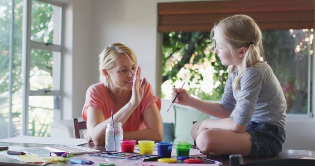 Happy caucasian mother with daughter painting pictures sitting on table in living room at home. Motherhood, childhood, creativity and togetherness.