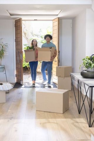 Young couple carrying moving boxes into bright modern home with houseplants and open wooden doors