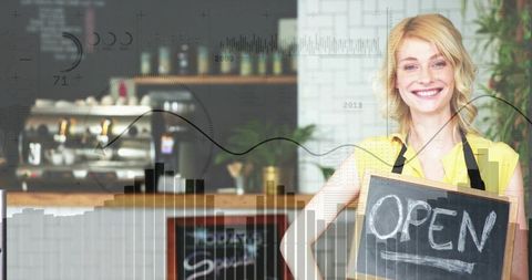 Smiling barista holding open sign at cafe counter with espresso machine and chalkboard