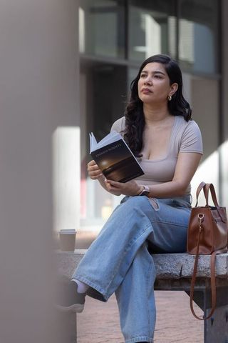 Young woman sitting on urban bench reading paperback, holding coffee, city lifestyle