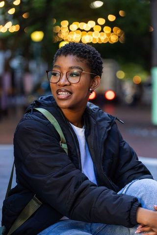 African woman sitting on bench at dusk wearing puffer jacket and glasses with city bokeh