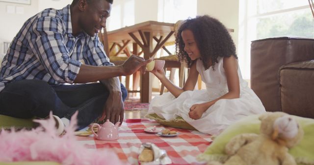 Father and daughter enjoying a tea party at home, sitting on a pink picnic blanket in the living room. This playful scene showcases family bonding, childhood imagination, and quality time together. Ideal for use in articles, blogs, advertisements, and campaigns related to family activities, parent-child relationships, and indoor entertainment.