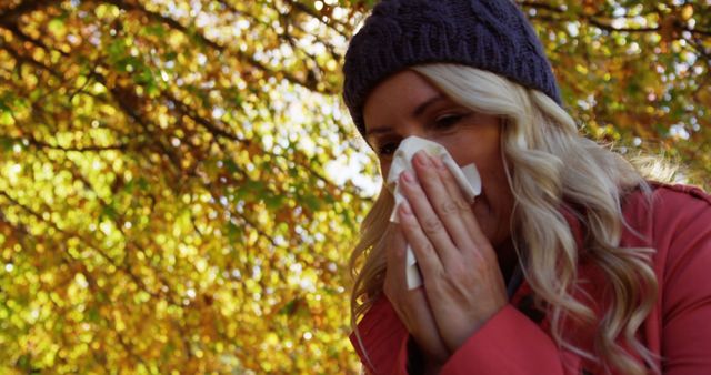 A woman with blonde hair is sneezing into a tissue while standing outdoors under a tree with colorful autumn leaves. She is wearing a hat and a pink coat, suggesting a cool, fall day. This image is ideal for use in content related to seasonal allergies, health and wellness, autumn activities, or outdoor life.