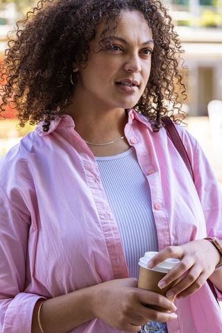Curly-haired woman holding coffee cup wearing pink shirt in sunny urban portrait