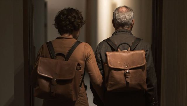 Couple walking down dim corridor wearing matching brown leather backpacks holding hands