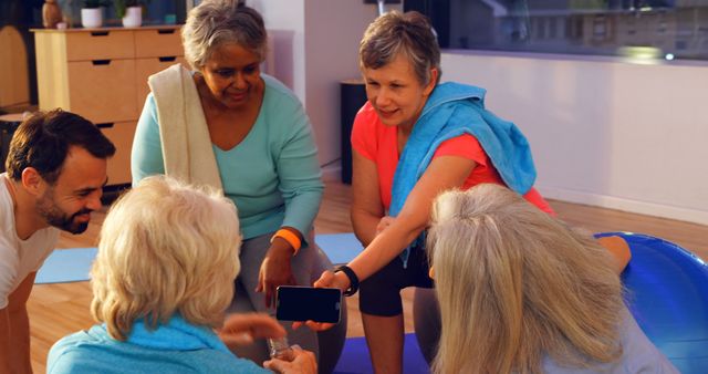 Senior individuals in fitness attire gather around a smartphone, likely sharing a fitness app or tracking progress. Blue and green exercise mats are visible, promoting health and wellness. Suitable for depicting senior fitness, technology use among elderly, community activities, group exercise, and promoting a healthy lifestyle for older adults.